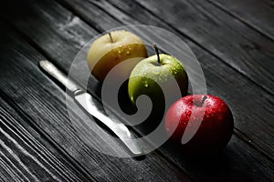 Red green yellow apples in a diagonal row with knife water drops on black wooden table, back light