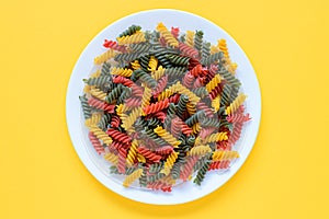 Red green and yellow dry raw fusilli pasta on a plate with yellow background.