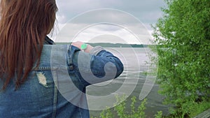 Red haird girl with green screened watches on hand looks at watches and picturasque scenry standing on old pier