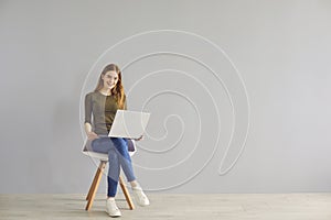 Red-haired beautiful girl student with a laptop sits on a chair on a gray background.