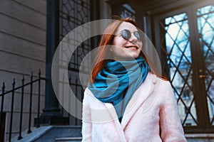 Red haired beautiful girl is walking by the street in a pink coat and blue scarf, with sunglasses.