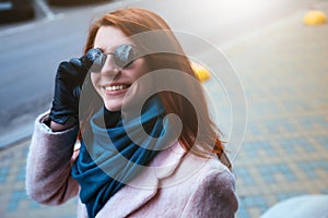 Red haired beautiful girl is walking by the street in a pink coat and blue scarf, with sunglasses.