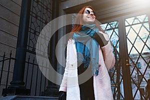 Red haired beautiful girl is walking by the street in a pink coat and blue scarf, with sunglasses.