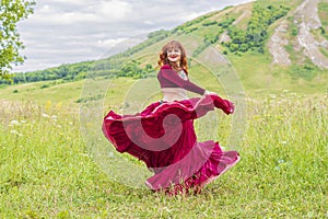 The red-haired beautiful young girl in a red dress is dancing a national dance on a green meadow.