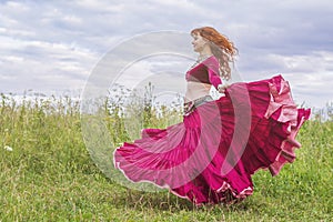 The red-haired beautiful young girl in a red dress is dancing a national dance on a green meadow.