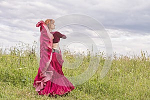 The red-haired beautiful young girl in a red dress is dancing a national dance on a green meadow.