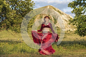 The red-haired beautiful young girl in a red dress is dancing a national dance on a green meadow.