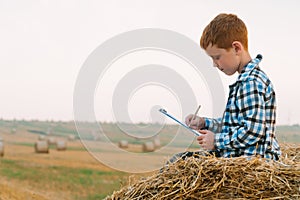 A red-haired boy on top of a straw bale holds a tablet with a letter and a pen in his hands on a background of hay bales
