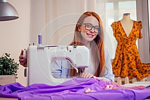 Red haired business woman with sewing machine and maniken in studio background. tailor creates a collection outfits sews