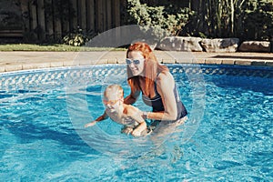 Red-haired Caucasian mother training baby son to swim in swimming pool outdoors. Baby boy in water goggles diving in water. Water