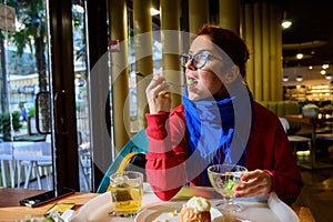 Red-haired caucasian woman eating salad in a cafe. Pensive romantic girl in glasses and a blue scarf is having lunch.