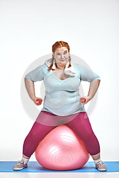 Red haired, chubby woman is sitting on the ball and keep the dumbbells