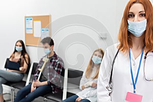 A red-haired doctor wearing a protective mask stands close to the camera in front of a queue in the lobby of an