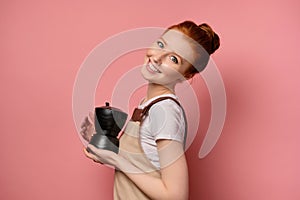 A red-haired girl in an apron stands in profile on a pink background with a coffee pot in hands and smiles broadly at