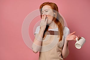 A red-haired girl in a apron and a T-shirt stands on a pink background, mocks, covering mouth with palm, turning the mug photo