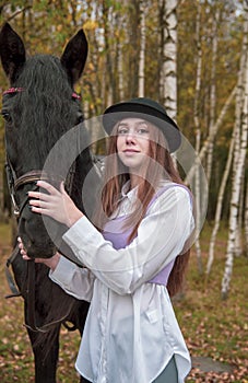 Red-haired girl in a black hat with a horse in the autumn forest.