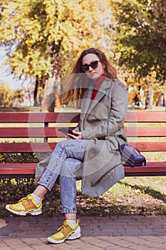 Red-haired girl in a coat sits on a red bench in autumn in the park