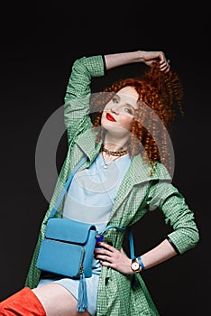 Red-haired girl, curly hair, holds hands on her head, pulls her hand with her hair, bright clothes, red lipstick,