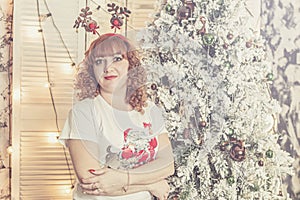 Red-haired girl in estive dress stands against backdrop of Christmas tree and looks into camera.