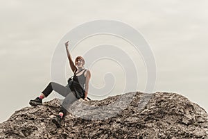 Red-haired girl gestures with a middle finger sitting on the stones above the precipice against the backdrop of mountains and