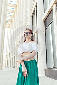 Red-haired girl in glasses stands against the background of a beautiful modern building