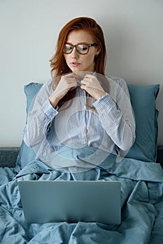 A red-haired girl with glasses zips up her shirt sitting in bed and looks into her laptop