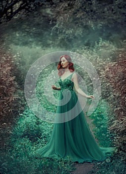 Red-haired girl in a green, emerald, luxurious dress in the floor, with a long train. The princess walks in a fairy