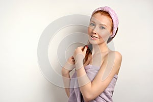 A red-haired girl in a lilac towel stands half-turned on a white background and looks at the camera, straightening hair