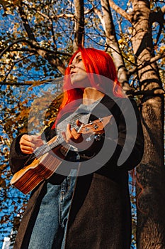 Red-haired girl with long hair plays on the ukulele in the park. School, music education concept, student learns to play the