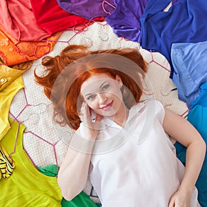 Red-haired girl is lying on the bed. Colorful bright clothes surround it.