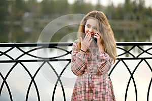 A red-haired girl in a pink checkered dress stands on the embankment and talks on a cell phone