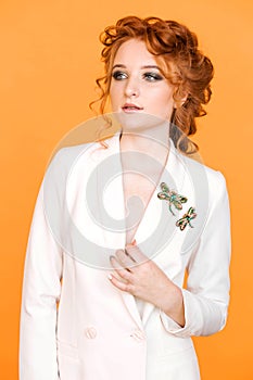 Red-haired girl posing on an orange background in a white jacket with a brooch in shape of dragonfly with green gemstone