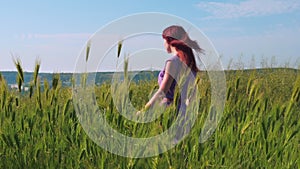 A red-haired girl in a purple dress walks along a path along a field of green grass and spikelets swaying in the wind.