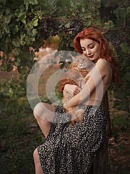 Red-haired girl with a red cat in the countryside near a vineyard on a stump.