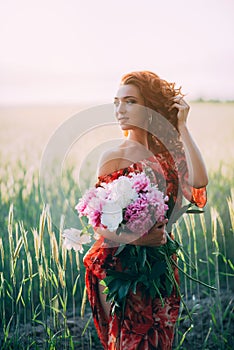 Red-haired girl in a red dress with a bouquet of peonies flowers field in summer at sunset. Woman`s happiness