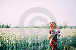 Red-haired girl in a red dress with a bouquet of peonies walking along a path in a summer field at sunset