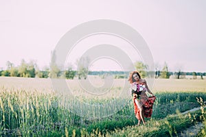 Red-haired girl in a red dress with a bouquet of peonies walking along a path in a summer field at sunset