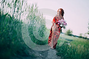 Red-haired girl in a red dress with a bouquet of peonies walking along a path in a summer field at sunset. Soft focus