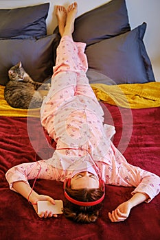 Red-haired girl in red pajamas on a red bed at home next to pet. A woman listens to music on headphones with a cat during a