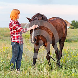 Red-haired girl in a red plaid shirt stroking a brown horse