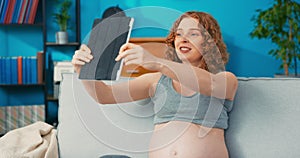 red-haired girl is relaxing on sofa in living room with tablet in hand, she is on maternity leave, video chatting online