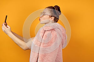 A red-haired girl in round glasses stands in profile in a pink coat and takes a selfie, stretching out lips in a kiss
