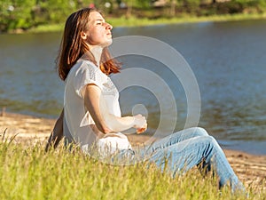 A red-haired girl sits in the grass by the lake and enjoys the evening sun during the sunset