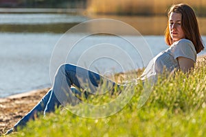 A red-haired girl sits in the grass by the lake and enjoys the evening sun during the sunset