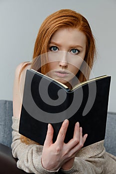 The red-haired girl sits leaning on the arm of the sofa and looks over the open black book in her hands.