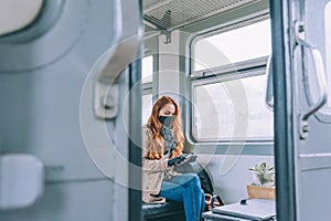 Red-haired girl sits in a train carriage, with a black medical mask, holding a smartphone in her hands.