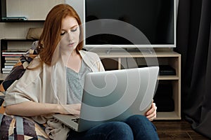 Red-haired girl sitting in a chair at home looking at a laptop on her lap