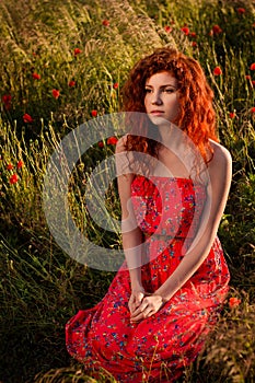 Red-haired girl sitting in the poppies meadow at sunset