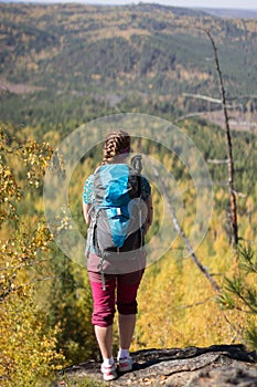 Red-haired girl standing with a backpack on a rock on a background of autumn forest