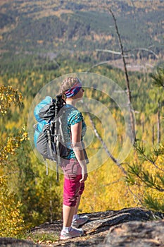 Red-haired girl standing with a backpack on a rock on a background of autumn forest